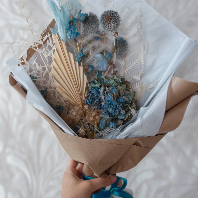 A small dried flower bouquet featuring blue and white flowers wrapped in paper with a blue crystal