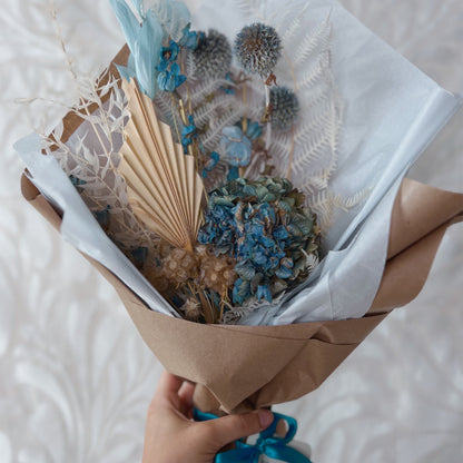 A small dried flower bouquet featuring blue and white flowers wrapped in paper with a blue crystal