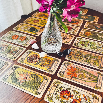 About a dozen of the Botanica Oculta Tarot Deck cards laid out flat, with a small vase of flowers and crystals nearby
