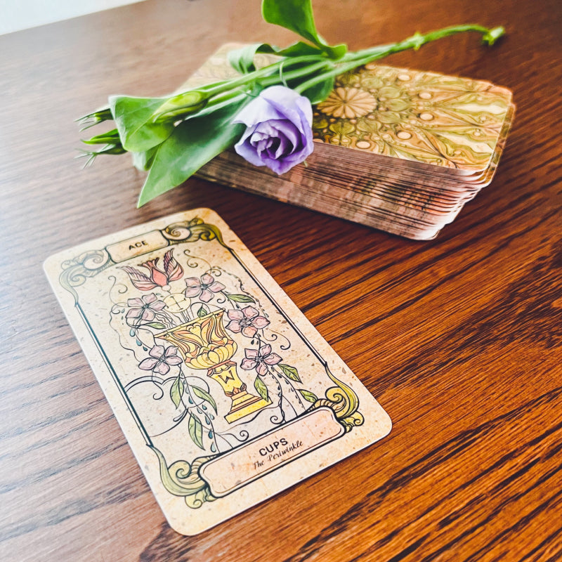 The Ace of Cups card from the Botanica Oculta Tarot Deck laid out on a wood table with a purple lisianthks flower