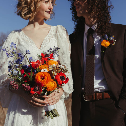 A bride and groom standing outside looking at each other smiling. He has a colorful boutonniere and she holds a colorful bouquet of jewel-toned flowers