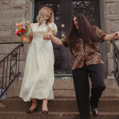Two brides, one in a white dress and the other in a cheetah print shirt and slacks, holding hands, laughing, walking down stairs