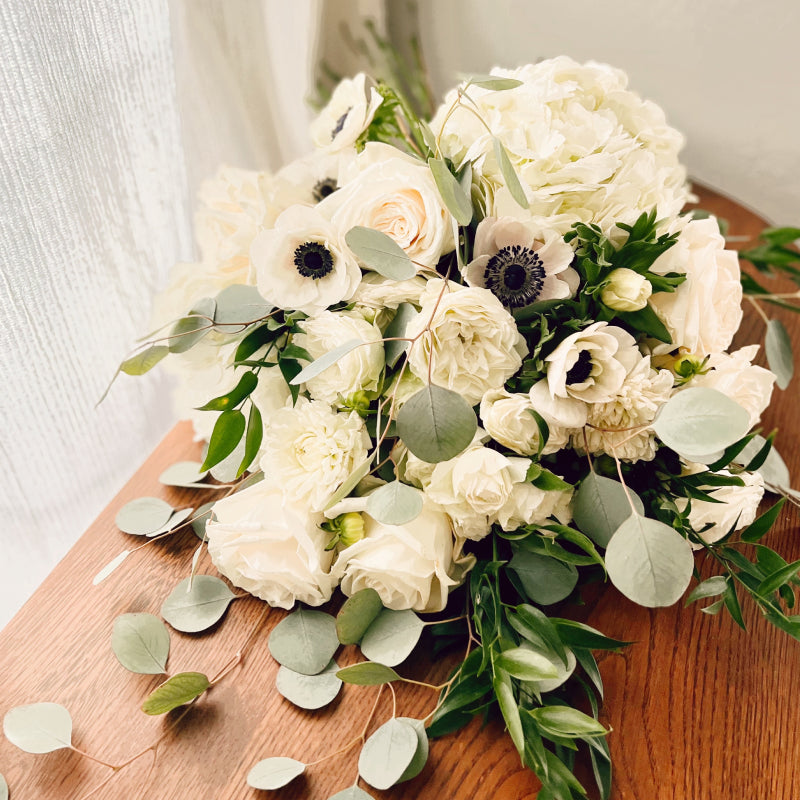 A neutral colored bridal bouquet featuring premium white flowers and eucalyptus, displayed laying on its side on a wooden table