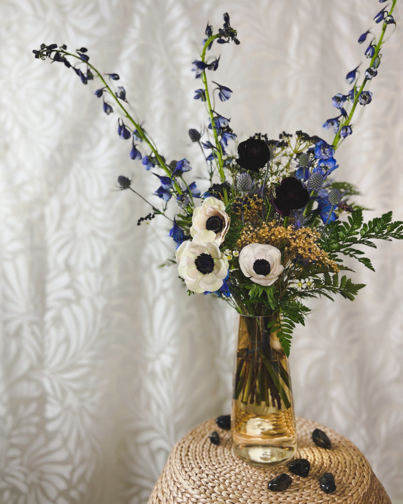 A bouquet of blue, white, black, and tan flowers displayed in an amber glass vase with blue tiger's eye crystals next to it
