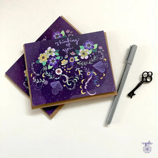 Dark purple greeting card with illustration of flowers, crystals, and moons that says "Thinking of You"