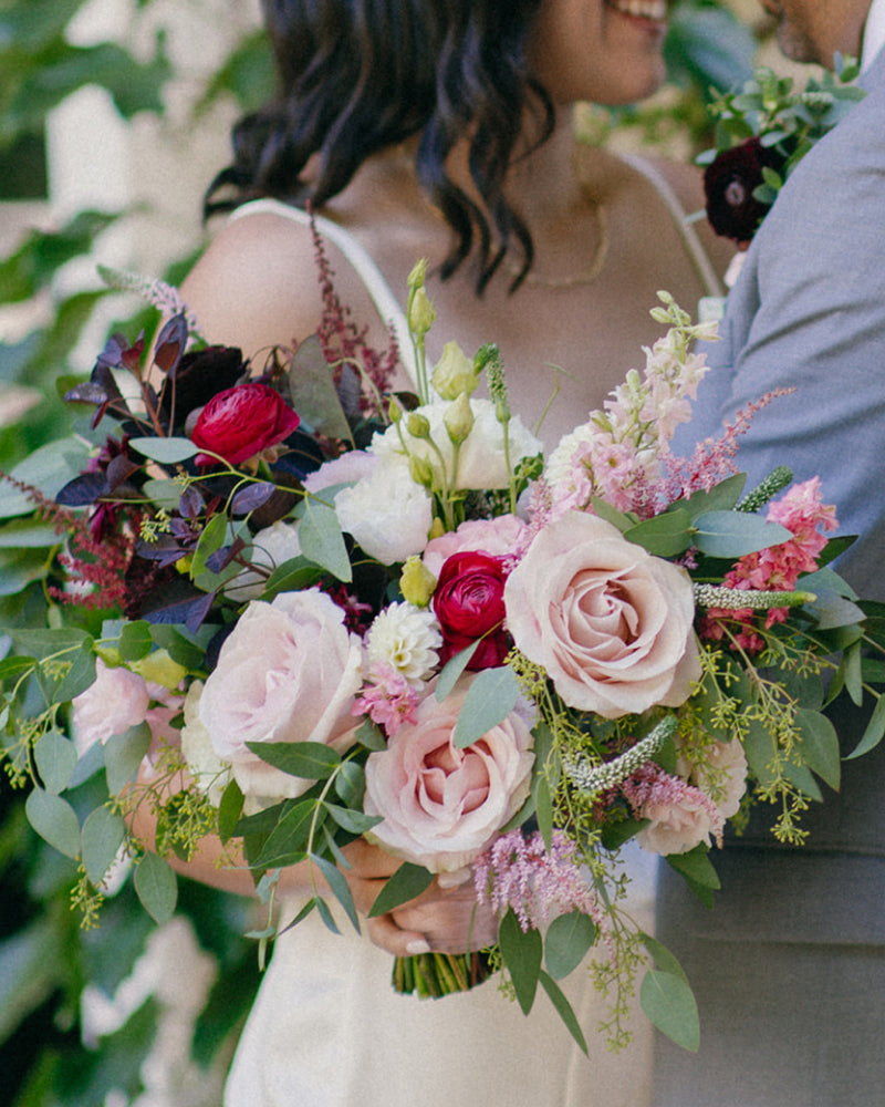 Up close view of a bride holding a bouquet of eucalyptus, blush roses, hot pink ranunculus, and more