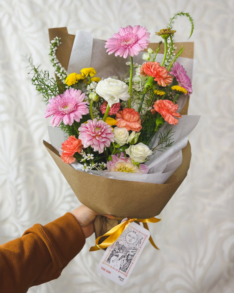 Hand holding a wrapped bouquet of warm, pastel flowers with a The Sun tarot card attached