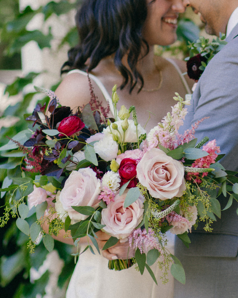 A close up shot of a bride holding out her green, pink, and maroon bouquet while she hugs her groom