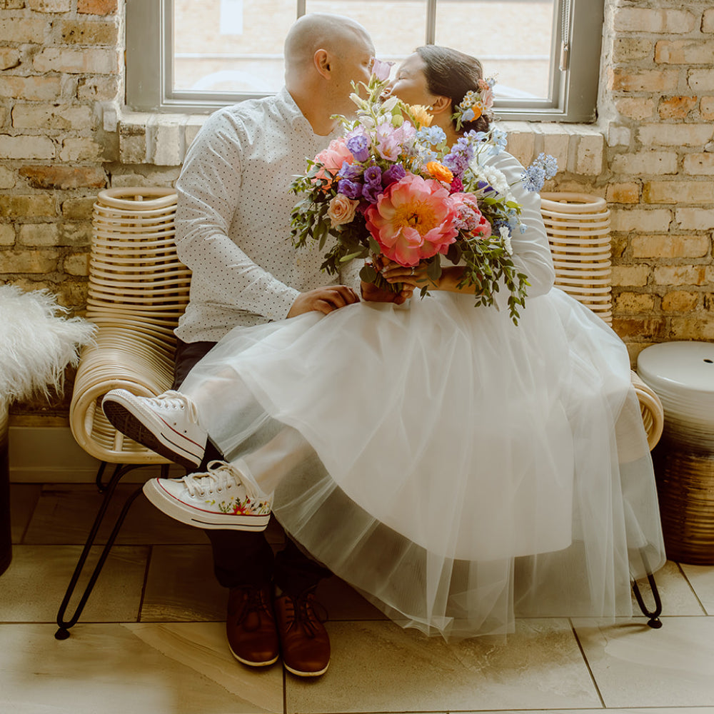 A bride sitting on a groom's lap kissing and holding a flower bouquet