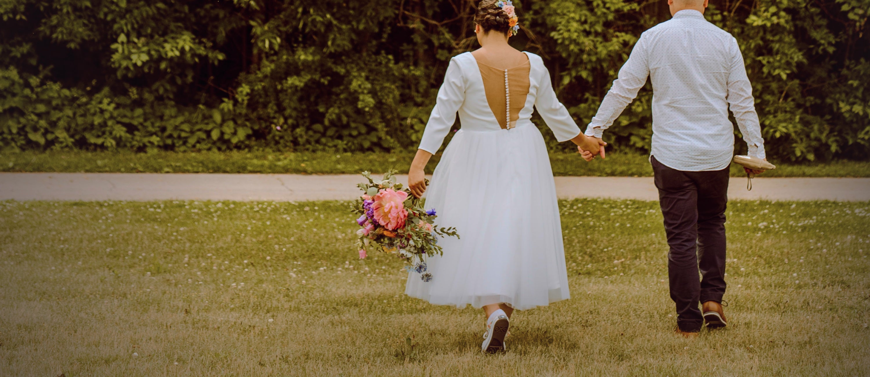 A bride and groom walking in the grass with their backs towards the candle. The bride is holding a colorful bouquet of flowers and wearing chucks.