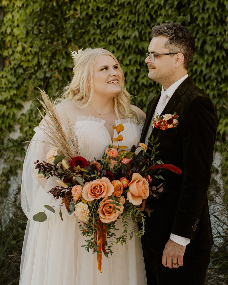 A bride and groom smiling at each other with a large bouquet of fall flowers