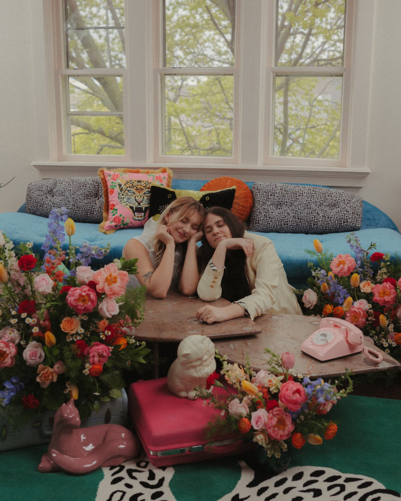 Two women setting on the floor in front of a couch surrounded by colorful fresh flowers and vintage decor