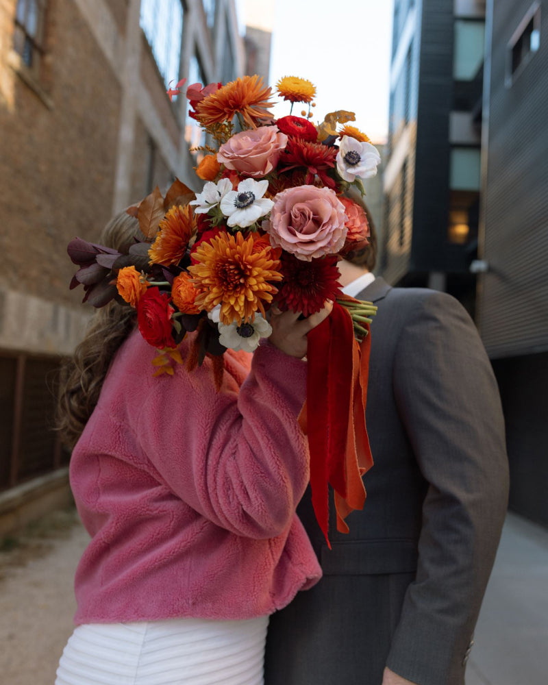 A bride in a pink jacket and her groom kissing while a bouquet covers their faces in a downtown city setting