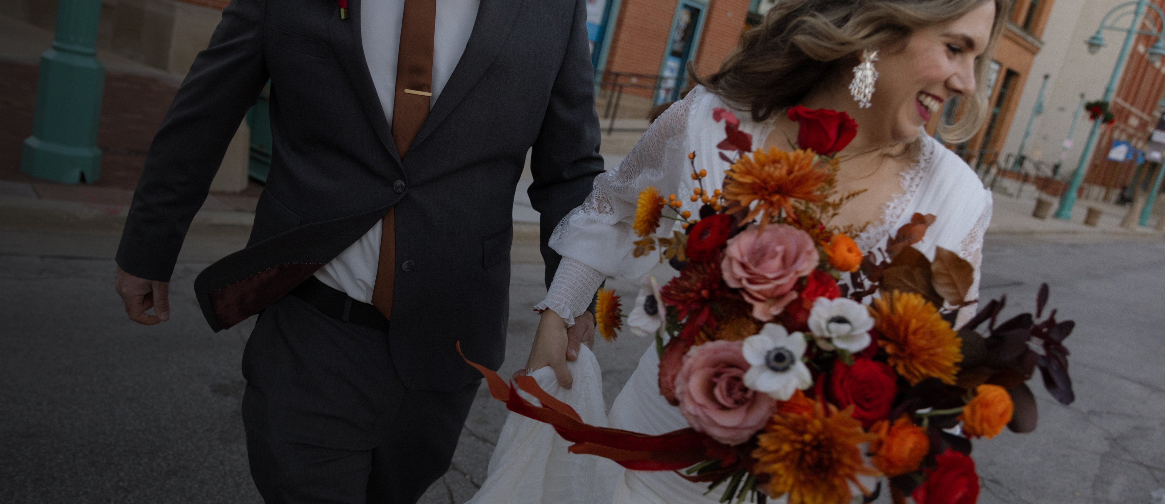 A bride running down a city street smiling, holding a large bouquet of flowers while her groom follows her