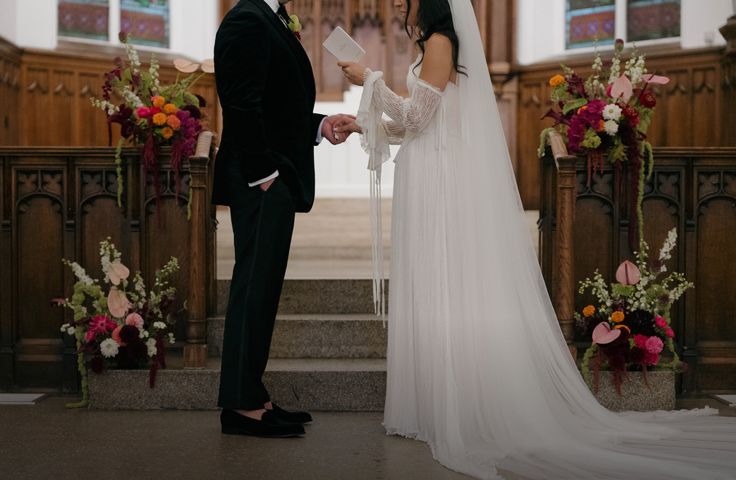 Bride holds a small book while reading vows to the groom at the altar. Four flower arrangements are behind them.