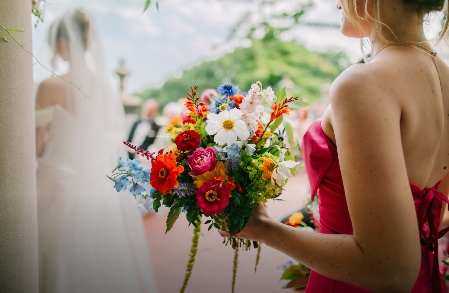 A bridesmaid in a pink dress holds the bride's colorful wedding bouquet, while the couple is married in the background.
