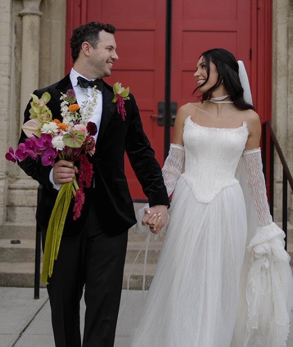 A bride and groom hold hands and look at each other, while the groom holds the bride's bouquet.