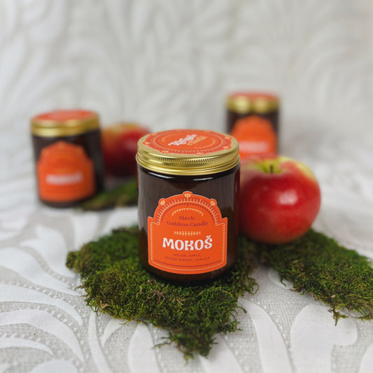 A candle in brown glass with an orange label that reads "Mokoś" displayed with moss and apples