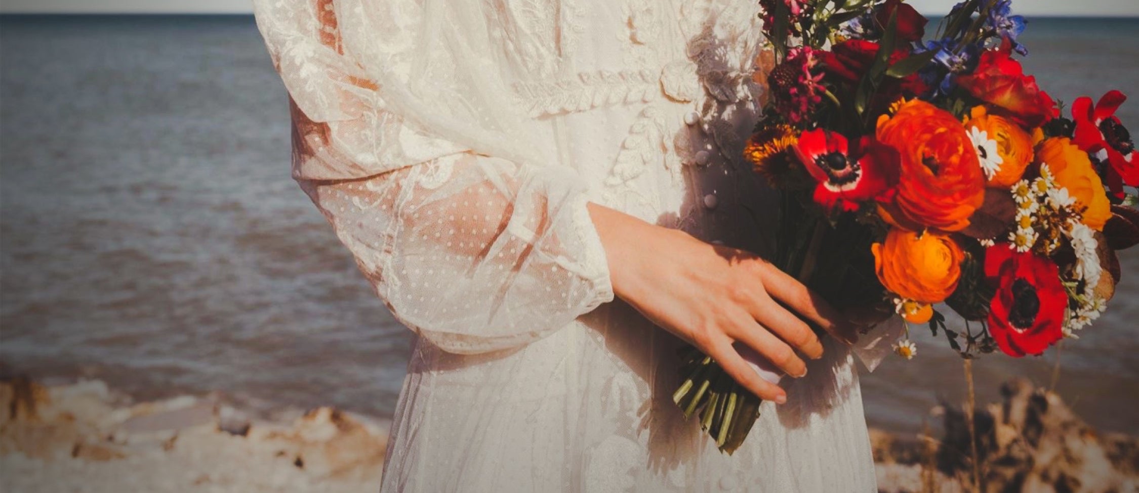 A close up of a bride in a long sleeved vintage lace dress holding a colorful bouquet of flowers with Lake Michigan in the background