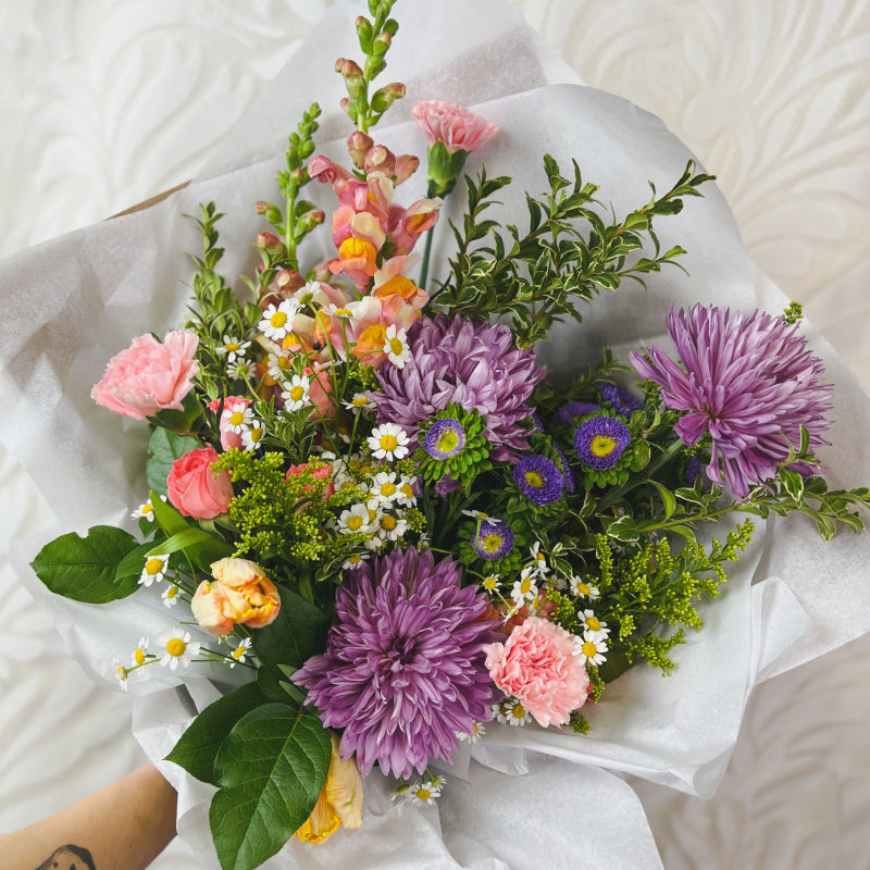 A colorful wrapped bouquet featuring light purple mums, pastel pink carnations, yellow tulips, and more
