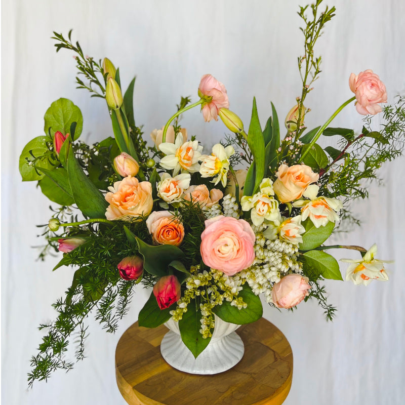 A springtime flower arrangement featuring warm colors in a white footed compote vase