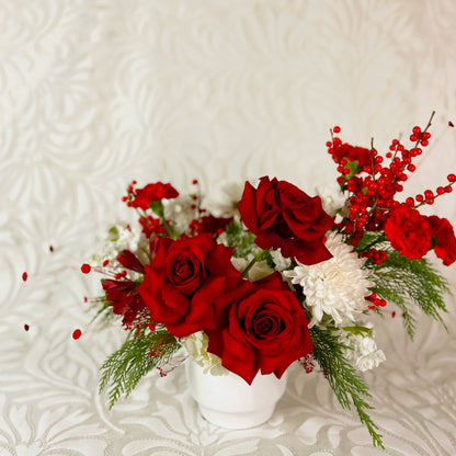 A holiday arrangement with red roses and berries, white mums, and evergreens in a white ceramic vase