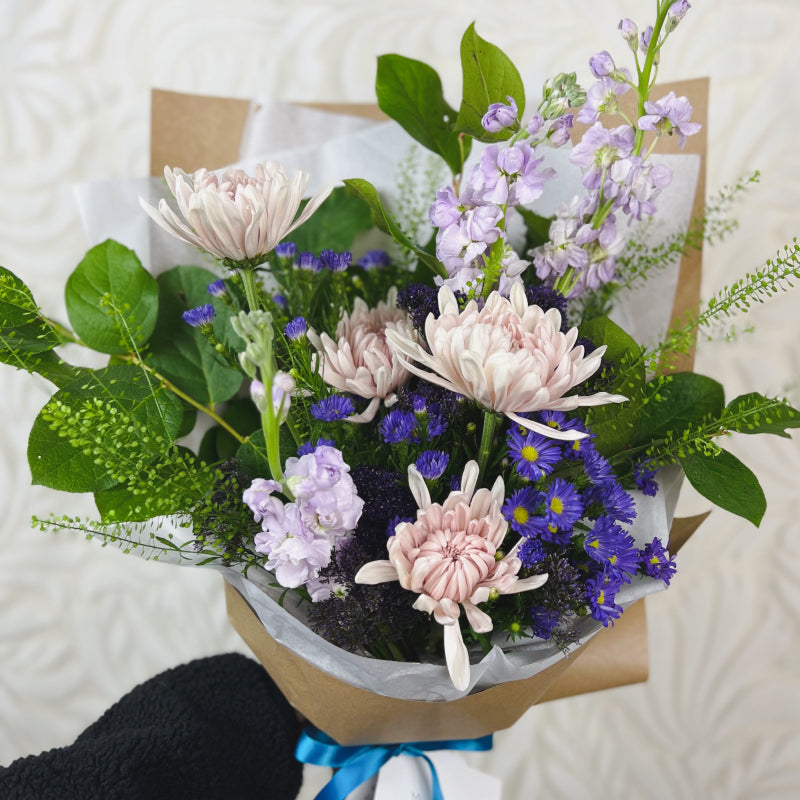 A small wrapped bouquet featuring greenery, light purple, and dark purple flowers