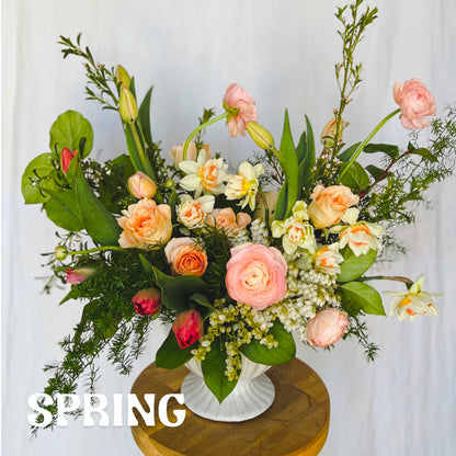 A large flower arrangement in a white footed compote vase featuring spring flowers like narcissus, tulips, and ranunculus. Text over image reads "Spring"