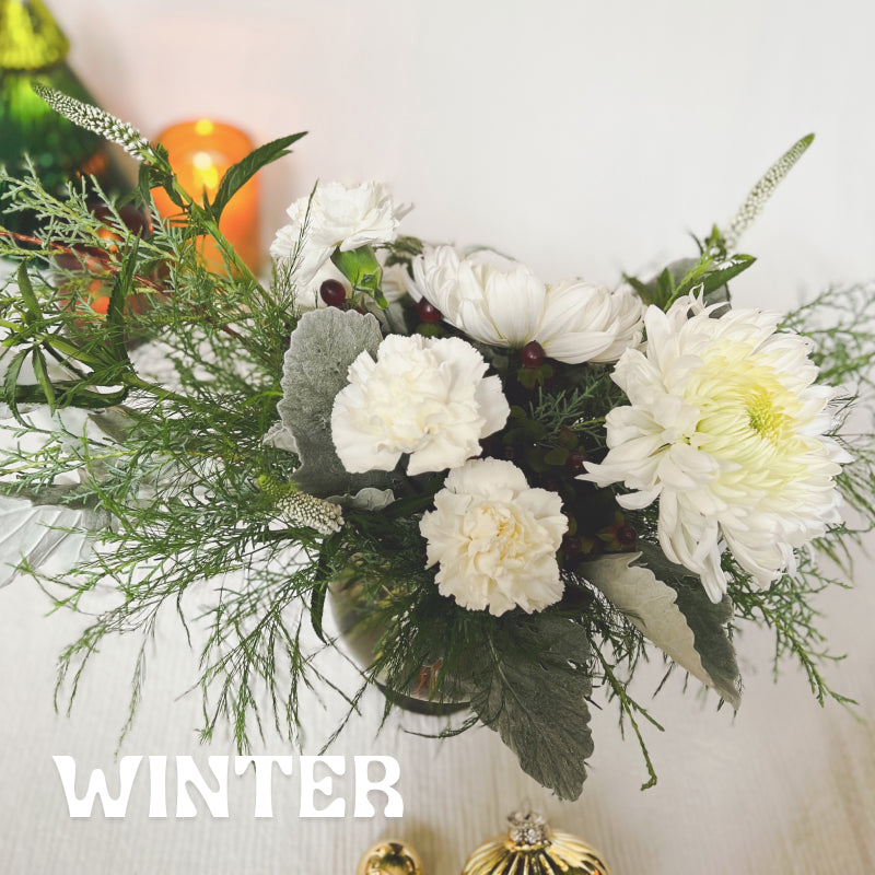 Evergreens, lambs ear, white carnations, white mums, and red berries in a vase, with holiday decorations visible in the background. Text reads "Winter."