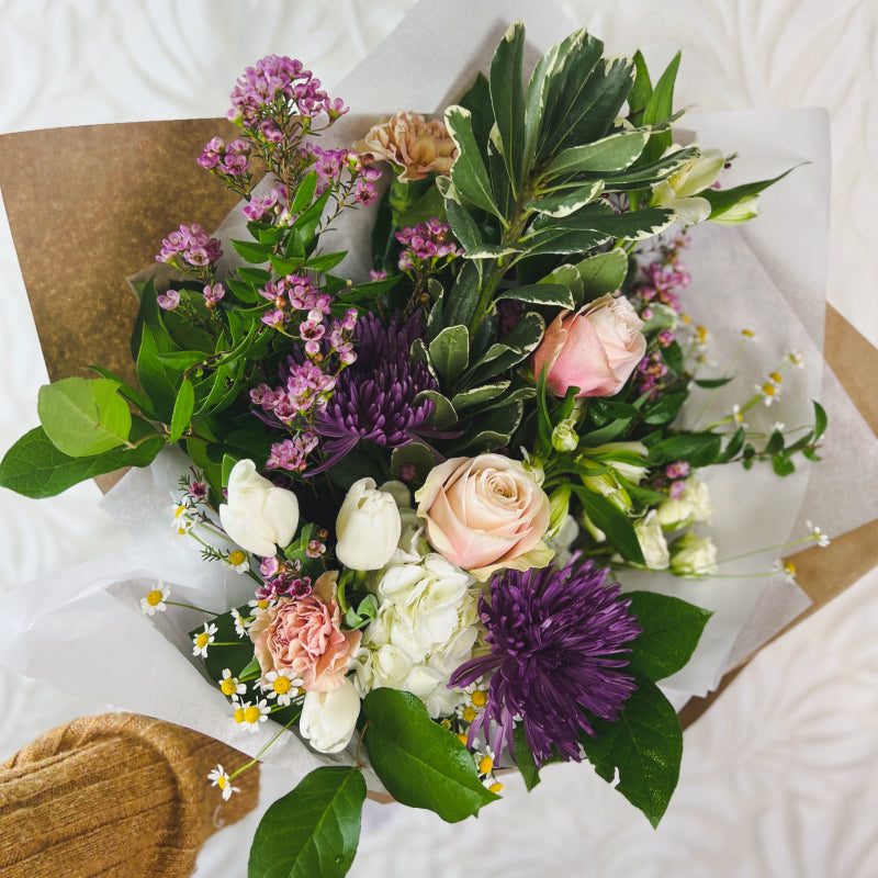 A wrapped fresh flower bouquet featuring pretty greenery, purple flowers, blush roses, and white tulips