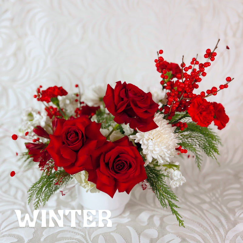 Red roses, red berries, white mums, and evergreens in a white vase. Text reads "Winter." 