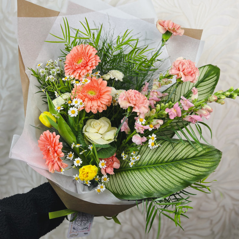 A wrapped flower bouquet featuring pink, peach, and yellow flowers with tropical greenery