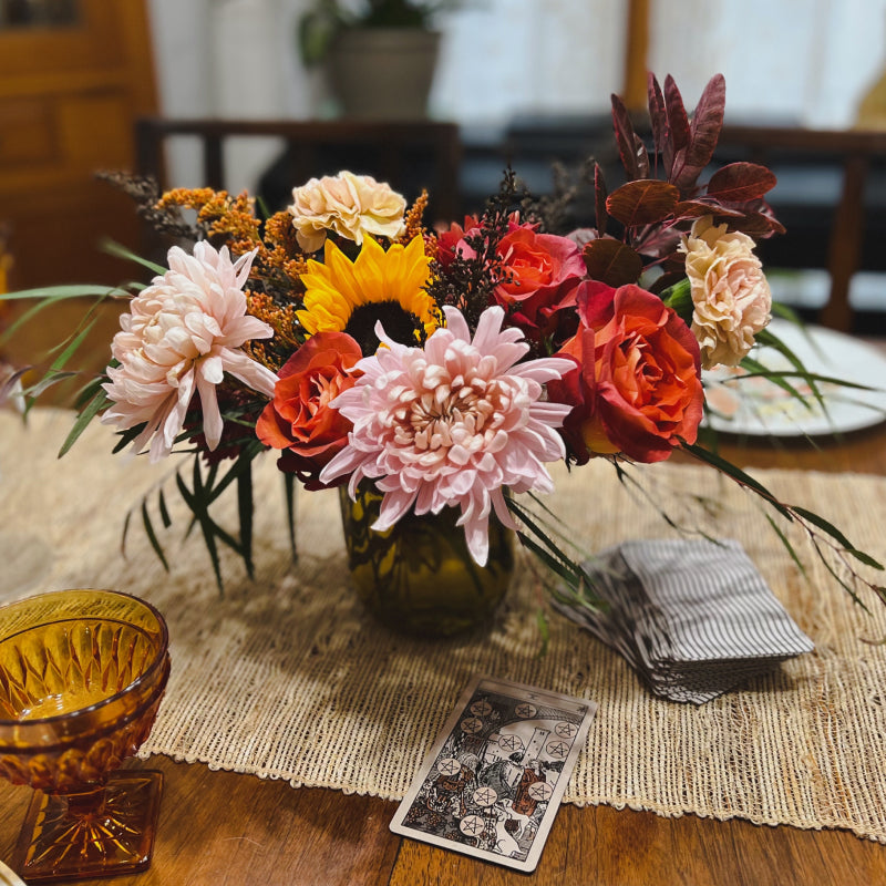 A small flower centerpiece featuring fall blooms with the ten of coins tarot card next to it