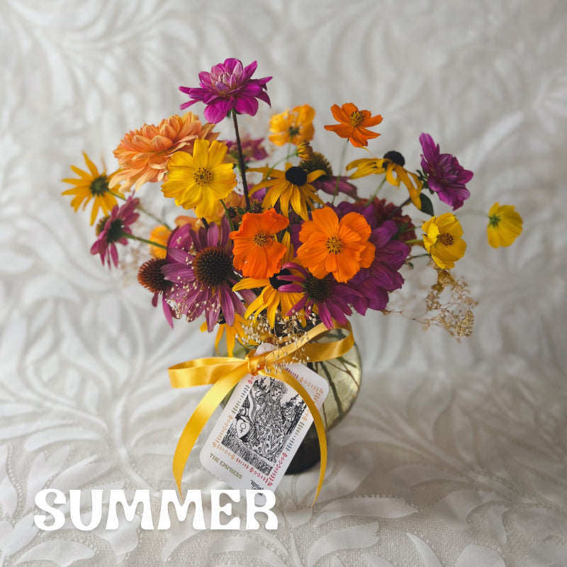 A small vase arrangement with bright yellow, orange, and purple flowers with a The Empress tarot card tied to it with ribbon. Text over image reads "Summer"