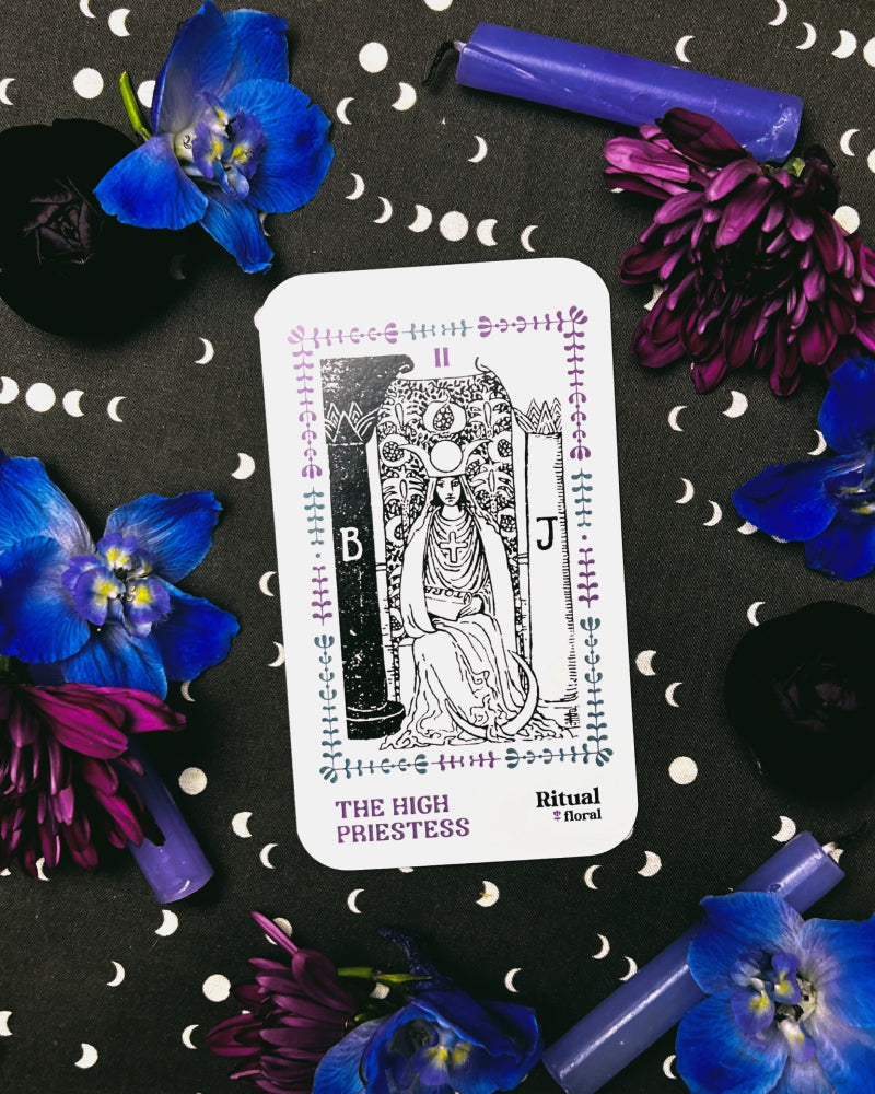 The High Priestess tarot card on a black background with blue and purple flowers and purple candles surrounding it