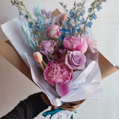 Wrapped bouquet of cool pastel flowers featuring peonies, tulips, roses, and more