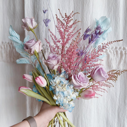 Hand holding a bouquet of flowers featuring cool, dreamy pastels like light blues, pinks, and purples