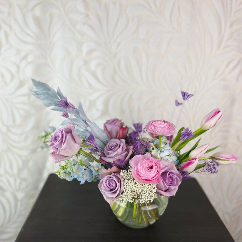 A short vase of fresh flowers in lavender, light blue, and pink.