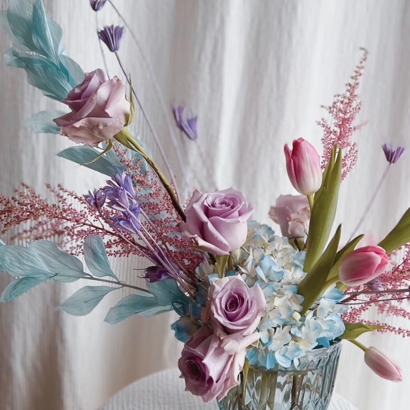 An up close shot of a flower arrangement, featuring a blue glass vase and a variety of cool pastel flowers, fresh and dried