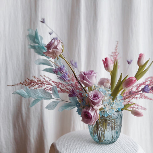 A dreamy flower arrangement in a blue glass vase featuring light blue, lavender, and pink pastel flowers