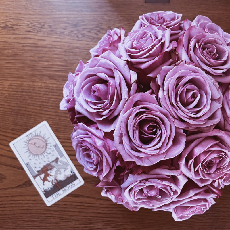 An arrangement of purple roses photographed from above with a The Moon tarot card