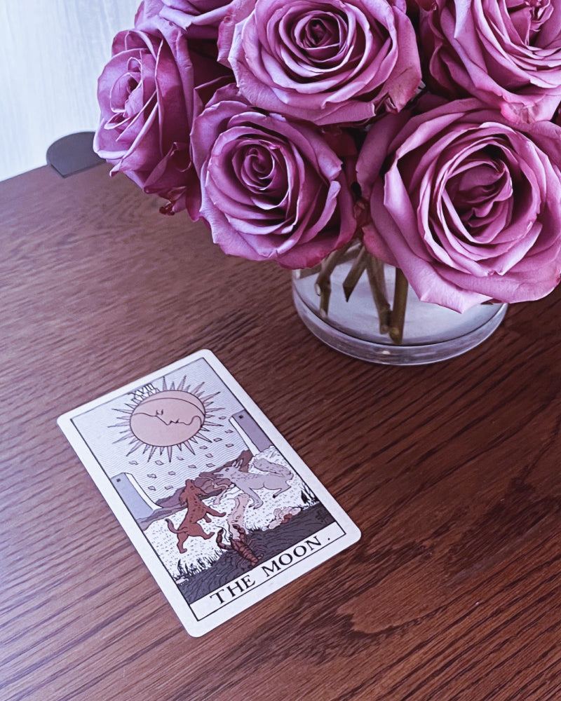 A The Moon tarot card next to an arrangement of purple roses
