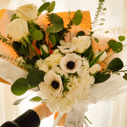 A wrapped flower bouquet featuring anemones, white roses, and eucalyptus