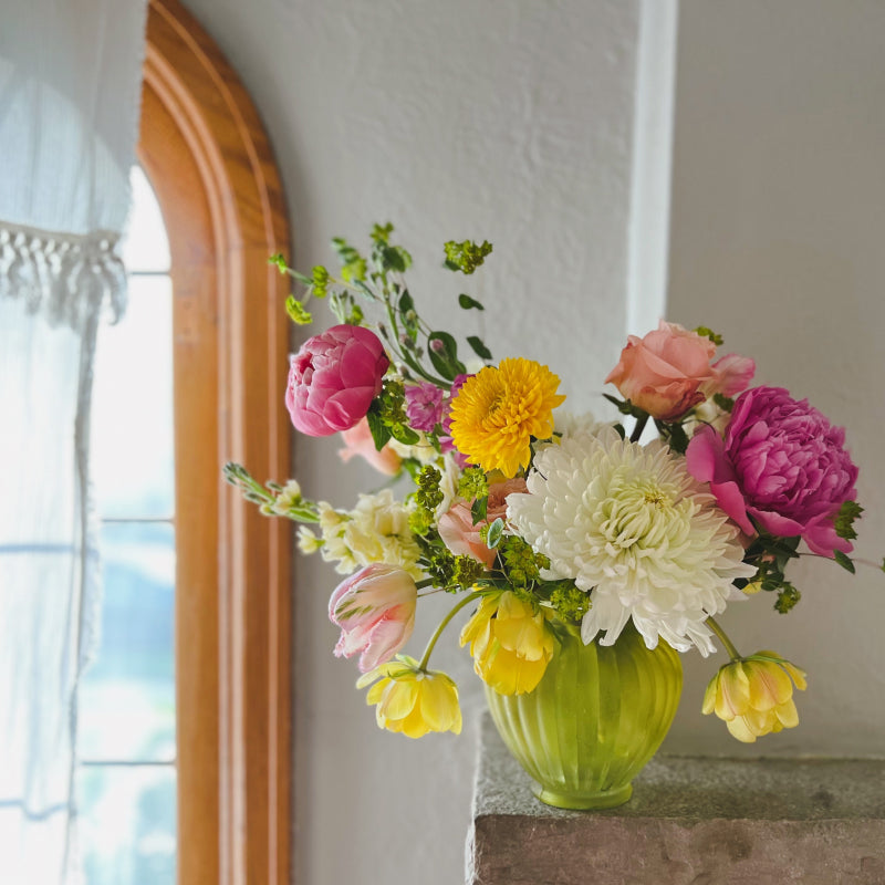 A vase arrangement of pink, yellow, and white flowers on a fire place mantel in front of a sunny window