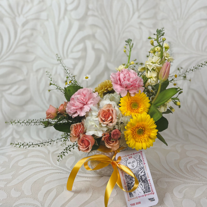 A small vase arrangement with warm pastel flowers and a The Sun tarot card tied to it with yellow ribbon