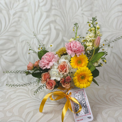 A small vase arrangement with warm pastel flowers and a The Sun tarot card tied to it with yellow ribbon