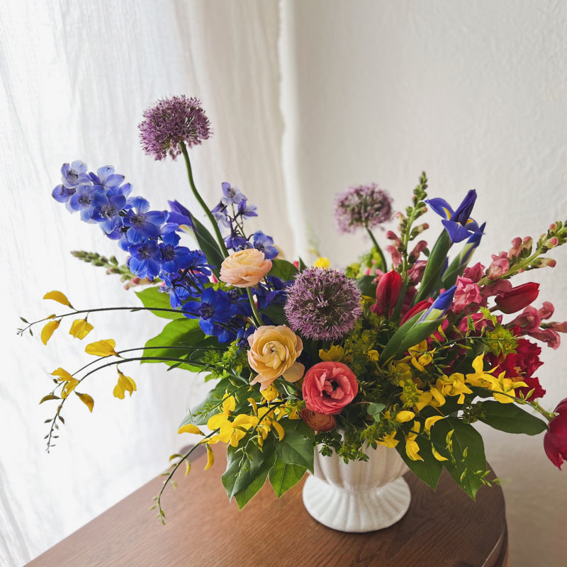 A bright and colorful arrangement of flowers in a white compote vase, featuring spring favorites like allium, ranunculus, iris, tulips, and more
