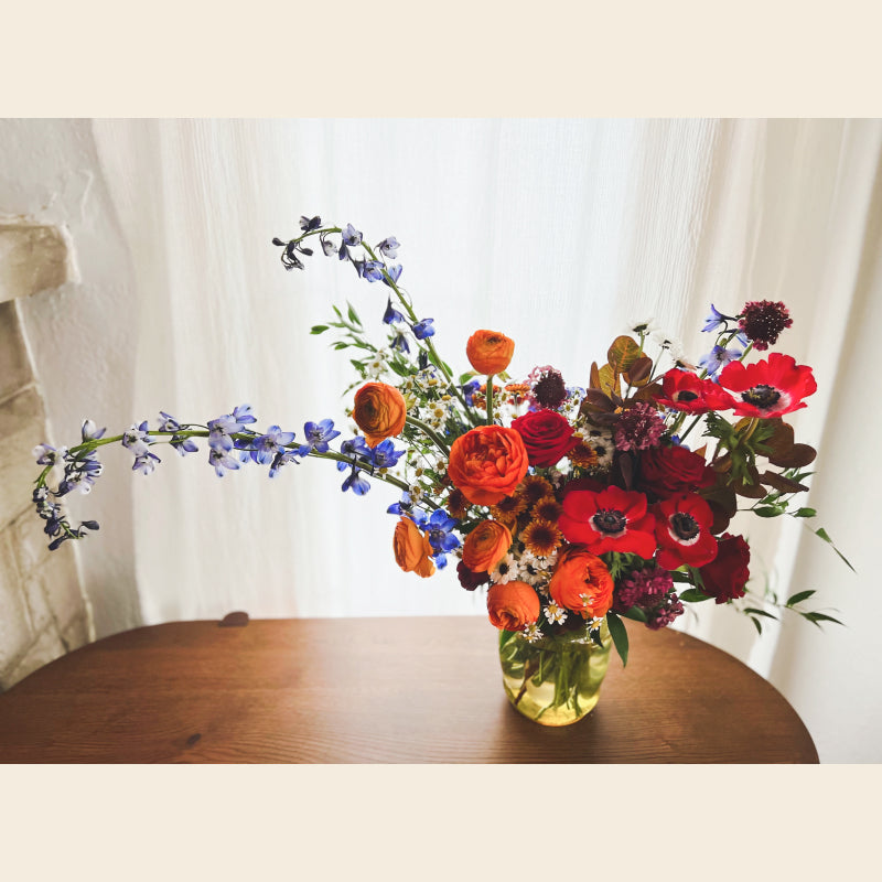 A lush, colorful arrangement of flowers featuring anemones, ranunculus, delphinium, and more