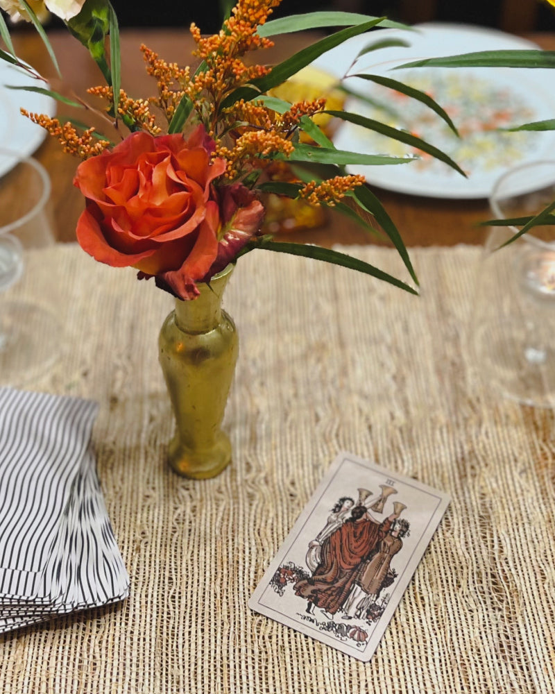 A gold bud vase of flowers and the 3 of cups tarot card on a dinner table