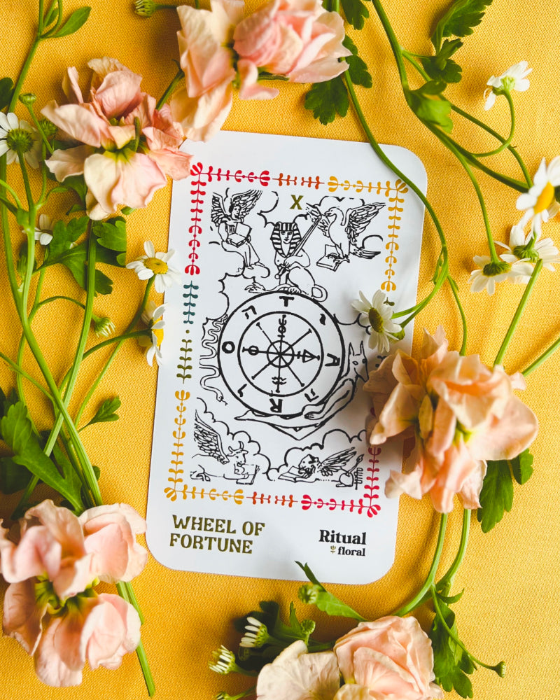 A Wheel of Fortune tarot card on a yellow background surrounded by real flowers